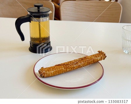 French press with herbal tea and rolled pastry on white table. Relaxed cafe atmosphere, cozy break, and warm daylight simplicity. French press with herbal tea and rolled pastry on white table. Relaxed cafe atmosphere, cozy break, and warm daylight simplicity. 133083317