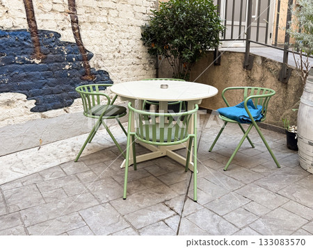 Round table and green chairs near wall in outdoor cafe. Cozy urban patio with plants, simplicity, and casual dining mood. 133083570