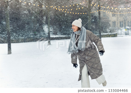 Portrait young adult smiling woman wear long quilted coat white hat enjoy play snowball snowy day outdoors. Snowflakes fall she moves playfully in front of stairs decorated with glowing winter lights 133084466