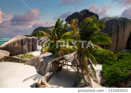 Granite boulders frame a narrow sandy path to turquoise water at left. Coconut palms and a thatched shelter sit on powder sand in warm late afternoon light. 133085003