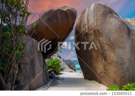 A narrow sandy path winds between granite boulders at Anse Source d'Argent, La Digue, Seychelles, framing turquoise waves and islands in warm late afternoon light. 133085011