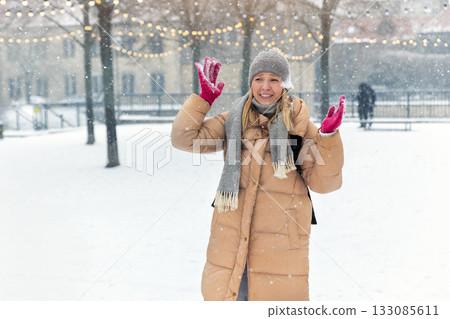 Portrait young adult smiling woman wear long quilted coat white hat enjoy play snowball snowy day outdoors. Snowflakes fall she moves playfully in front of stairs decorated with glowing winter lights Portrait young adult smiling woman wear long quilted coat white hat enjoy play snowball snowy day outdoors. Snowflakes fall she moves playfully in front of stairs decorated with glowing winter lights 133085611