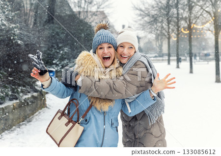 Portrait two happy women enjoy snowy winter day smiling hugging each other street covered fresh snow. Warm coats knitted hats scarves emphasize joyful cold season mood friendship festive atmosphere Portrait two happy women enjoy snowy winter day smiling hugging each other street covered fresh snow. Warm coats knitted hats scarves emphasize joyful cold season mood friendship festive atmosphere 133085612