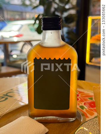 Glass bottle with amber liquid on wooden cafe table. Beverage photography, lighting reflection, and minimal composition with bokeh. 133085662