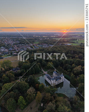 Aerial view at sunset white castle like building with moat and towers nestled in dense forest, winding path leads to entrance, warm sky glows over nearby town blending history, nature, and modern life 133085741