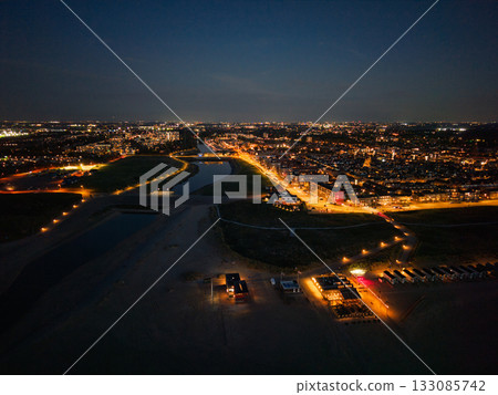 Aerial nighttime view of coastal city lit beach huts and restaurant foreground canal flanked by green zones and dense urban lights, blending natural darkness with vibrant infrastructure glow. Aerial nighttime view of coastal city lit beach huts and restaurant foreground canal flanked by green zones and dense urban lights, blending natural darkness with vibrant infrastructure glow. 133085742