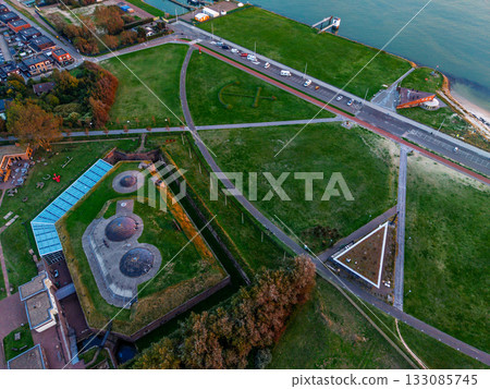 Aerial view of coastal urban recreational zone road with vehicles, red bike path, circular lookout, geometric green fields, tennis courts, and dense residential blocks framed by forest and shoreline. 133085745