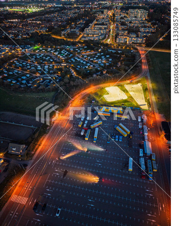 Aerial night view of Circus Renz setup bright skatepark and striped tents glow beside organized bus filled parking lot, residential grid and warm streetlights frame lively recreational zone. 133085749