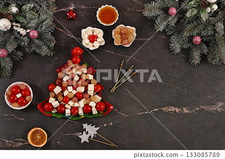 Festive New Year's tree-shaped salad with feta cheese, tomatoes, grapes, olives, and honey, topped with fir branches and decorations. Dish design idea. Christmas and New Year's dishes, menu template Festive New Year's tree-shaped salad with feta cheese, tomatoes, grapes, olives, and honey, topped with fir branches and decorations. Dish design idea. Christmas and New Year's dishes, menu template 133085789