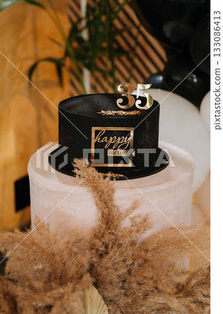 Black birthday cake with gold 35 topper on pedestal with pampas decor at elegant party Black birthday cake with gold 35 topper on pedestal with pampas decor at elegant party 133086413