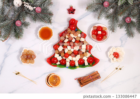 Festive New Year's tree-shaped salad with feta cheese, tomatoes, grapes, olives, and honey, topped with fir branches and decorations. Dish design idea. Christmas and New Year's dishes, menu template Festive New Year's tree-shaped salad with feta cheese, tomatoes, grapes, olives, and honey, topped with fir branches and decorations. Dish design idea. Christmas and New Year's dishes, menu template 133086447