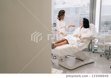 Remote view of woman client in bathrobe attentively listening to cosmetologist during in-clinic consultation about personalized aesthetic treatments, skin care plans and wellness options. Remote view of woman client in bathrobe attentively listening to cosmetologist during in-clinic consultation about personalized aesthetic treatments, skin care plans and wellness options. 133086649