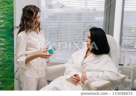 Cosmetologist showing product to client sitting in white robe, discussing personalized skincare solutions during professional consultation for health and beauty in contemporary clinic. Cosmetologist showing product to client sitting in white robe, discussing personalized skincare solutions during professional consultation for health and beauty in contemporary clinic. 133086655