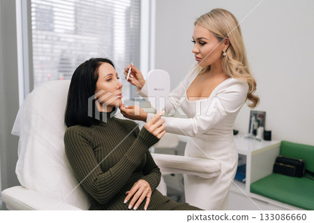 Female client looking in mirror while cosmetologist marking face for aesthetic procedure, preparing for beauty treatment in cosmetology clinic. Concept of facial anti-aging, skincare treatment. Female client looking in mirror while cosmetologist marking face for aesthetic procedure, preparing for beauty treatment in cosmetology clinic. Concept of facial anti-aging, skincare treatment. 133086660