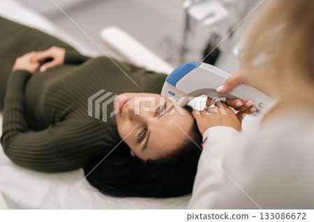 Portrait of middle-aged woman getting professional aesthetic ultrasound lifting procedure on forehead by cosmetologist in clinic. Aiming for skin rejuvenation and wrinkle reduction. 133086672