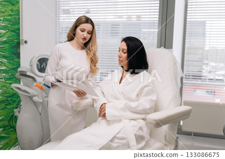 Aesthetician doctor consulting with client woman in bathrobe at beauty clinic, discussing treatments while reviewing document for skincare, wellness services. Concept of facial anti-aging treatment. Aesthetician doctor consulting with client woman in bathrobe at beauty clinic, discussing treatments while reviewing document for skincare, wellness services. Concept of facial anti-aging treatment. 133086675