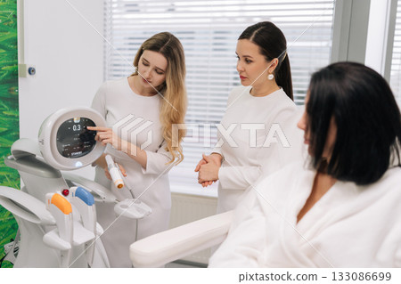 Cosmetologist female explaining settings on modern aesthetic device to client in white robe while another professional observes, offering consultation in bright modern aesthetic clinic. Cosmetologist female explaining settings on modern aesthetic device to client in white robe while another professional observes, offering consultation in bright modern aesthetic clinic. 133086699