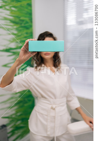 Young woman ophthalmologist in white coat holding a light-blue product box over her eyes in a modern clinic, symbolizing eye care, vision solution advertising and medical wellness Young woman ophthalmologist in white coat holding a light-blue product box over her eyes in a modern clinic, symbolizing eye care, vision solution advertising and medical wellness 133086700