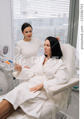 Cosmetologist consulting middle-aged client in modern beauty clinic, discussing skin care and aesthetic treatments, client examining her appearance in handheld mirror during wellness session. Cosmetologist consulting middle-aged client in modern beauty clinic, discussing skin care and aesthetic treatments, client examining her appearance in handheld mirror during wellness session. 133086703