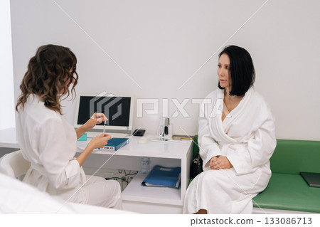 Skilled cosmetologist preparing syringe for cosmetic injection consulting with middle-aged female patient in bathrobe, focusing on beauty, facial anti-aging, skin care treatment in medical office. Skilled cosmetologist preparing syringe for cosmetic injection consulting with middle-aged female patient in bathrobe, focusing on beauty, facial anti-aging, skin care treatment in medical office. 133086713