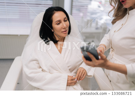 Close-up of beautician doctor in white uniform reviewing skincare treatment options on smartphone with female client in bathrobe during professional medical aesthetic consultation in clinic. Close-up of beautician doctor in white uniform reviewing skincare treatment options on smartphone with female client in bathrobe during professional medical aesthetic consultation in clinic. 133086721
