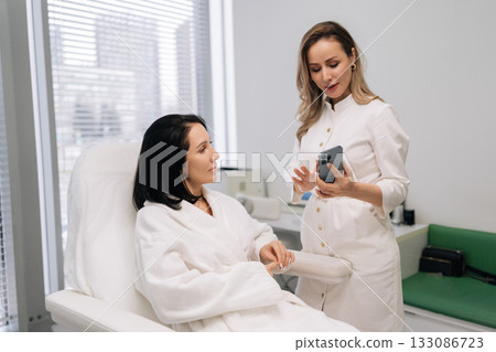 Aesthetician doctor in white uniform reviewing skincare treatment options on smartphone with female client in bathrobe during professional medical aesthetic consultation in clinic. Aesthetician doctor in white uniform reviewing skincare treatment options on smartphone with female client in bathrobe during professional medical aesthetic consultation in clinic. 133086723