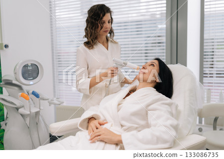 Professional cosmetologist performing ultrasound facial lifting treatment on woman face in beauty clinic, focusing on skin rejuvenation and anti-aging care. Concept of facial antiaging treatment. 133086735