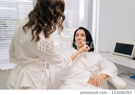 Rear view of experienced beauty technician consulting with woman seated comfortably in chair, using pencil to outline features for upcoming cosmetic enhancement at contemporary beauty center. 133086763