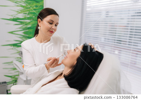 Side view of female client preparing for cosmetic treatment, medical surgeon marking face for aesthetic procedures in cosmetology clinic. Concept of facial anti-aging, skincare treatment. Side view of female client preparing for cosmetic treatment, medical surgeon marking face for aesthetic procedures in cosmetology clinic. Concept of facial anti-aging, skincare treatment. 133086764