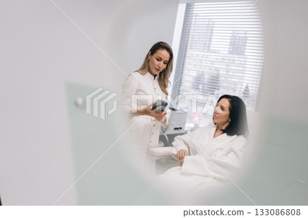 Reflection in mirror of aesthetician doctor consulting middle-aged female client in beauty clinic, showing information on mobile phone for skincare treatment planning in modern medical spa office. 133086808