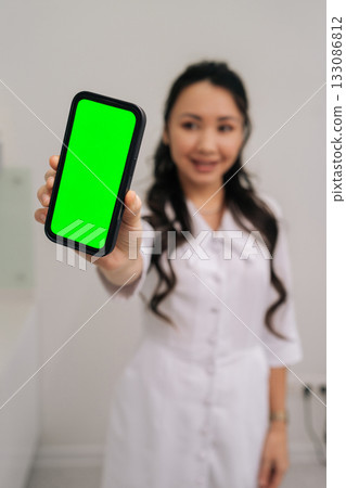 Smiling Asian cosmetologist presenting smartphone with green screen for mobile application display, offering mockups for product showcases, digital healthcare services, and beauty industry marketing. Smiling Asian cosmetologist presenting smartphone with green screen for mobile application display, offering mockups for product showcases, digital healthcare services, and beauty industry marketing. 133086812