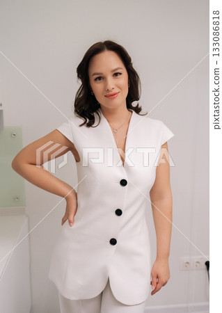 Vertical portrait of female cosmetologist smiling looking at camera, wearing uniform and conveying professionalism, expertise and caring patient-focused aesthetic medicine service in beauty clinic. 133086818
