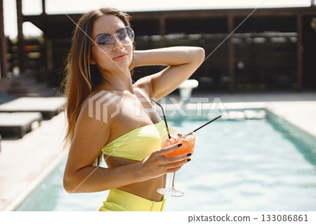 Woman in swimwear holding glass and drinking cocktail near swimming pool. Woman in swimwear holding glass and drinking cocktail near swimming pool. 133086861