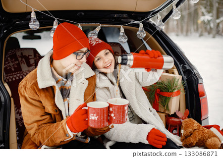 Joyful teens brother and sister spending time together in the winter park. Boy and girl sitting in open car's trunk and holding thermos and red cups with tea. Family wearing warm jackets and knitted Joyful teens brother and sister spending time together in the winter park. Boy and girl sitting in open car's trunk and holding thermos and red cups with tea. Family wearing warm jackets and knitted 133086871