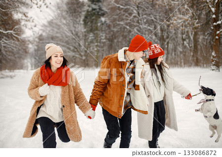 Joyful teens, their beautiful mother and black dog spending time together in the winter park. Family runing on a snow. Family wearing warm jackets and knitted hats. 133086872