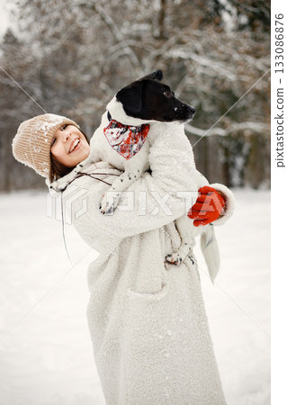Girl spending time in the winter park with her black dog. Teenager posing for a photo. Girl wearing warm jacket and knitted hat. Girl spending time in the winter park with her black dog. Teenager posing for a photo. Girl wearing warm jacket and knitted hat. 133086876