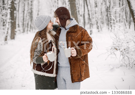 Bearded man and brunette woman in forest on snow day. Wife and husband wearing warm clothes. Couple hugging and holding two cups of cocoa drink with marshmallow and candy hook. 133086888