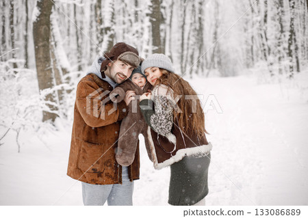 Parents with their baby son having fun in forest on snow day. Bearded man, brunette woman and baby son wearing warm clothes. Mother and father holding their son on hands. 133086889