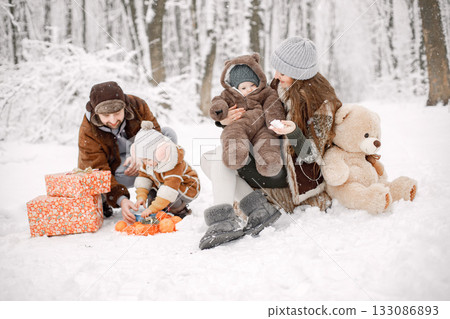 Parents with their children having fun in forest on snow day. Happy toddler daughter and baby son wearing warm clothes. Gift boxes standing next to them. Parents with their children having fun in forest on snow day. Happy toddler daughter and baby son wearing warm clothes. Gift boxes standing next to them. 133086893