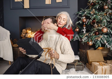 Senior man celebrating christmas with his wife near beautiful christmas tree. Old bearded man sitting on a floor near his wife with grey hair and holding a gifts. Man wearing white sweater and woman 133086905