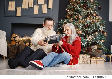 Senior man celebrating christmas with his wife near beautiful christmas tree. Old bearded man sitting on a floor near his wife with grey hair and holding a gifts. Man wearing white sweater and woman Senior man celebrating christmas with his wife near beautiful christmas tree. Old bearded man sitting on a floor near his wife with grey hair and holding a gifts. Man wearing white sweater and woman 133086906