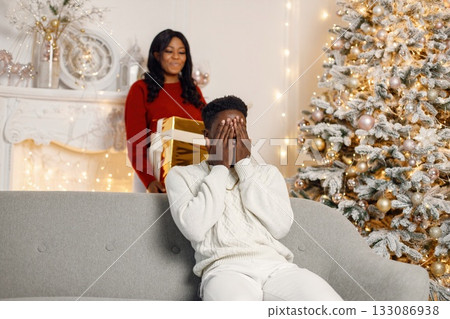 Young black couple with gifts in Christmas eve. Man sitting on a coach at home. Brunette woman standing behind and presented a gift for her boyfriend. Young black couple with gifts in Christmas eve. Man sitting on a coach at home. Brunette woman standing behind and presented a gift for her boyfriend. 133086938