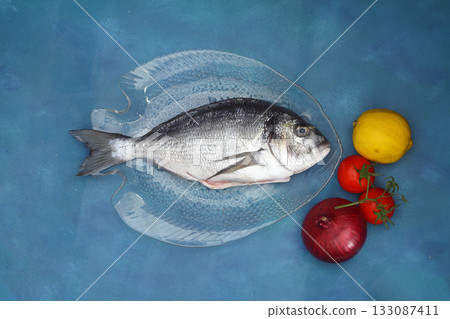 fresh fish on a blue wooden board. Raw uncooked sea bream on glass plate. With onion, lemon and tomatos 133087411