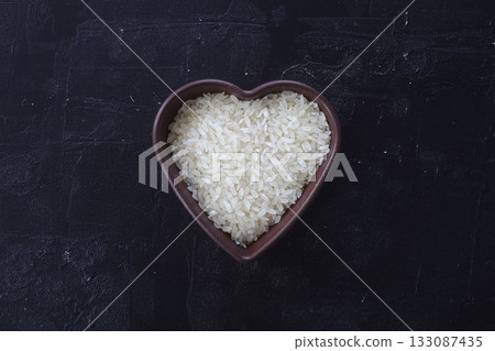 Isolated handful of raw white rice in the wood bowl in the shape of heart on black stone background 133087435