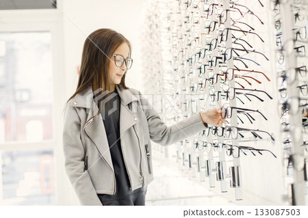 Little girl choosing eyeglasses in optica shop. Female customer taking eyeglasses from shelves in optica. Girl wants to try eyeglasses on. Little girl choosing eyeglasses in optica shop. Female customer taking eyeglasses from shelves in optica. Girl wants to try eyeglasses on. 133087503