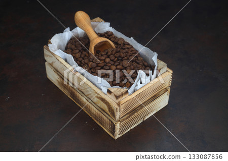 Closeup of roasted coffee beans in a small wooden crate, isolated on dark background. With wooden spoon. Closeup of roasted coffee beans in a small wooden crate, isolated on dark background. With wooden spoon. 133087856