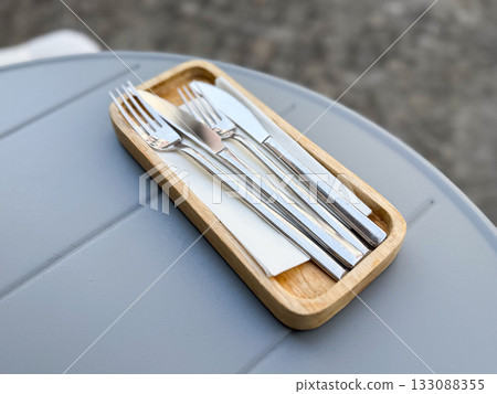 Wooden tray with metal cutlery and napkin on gray table. Dining setup, minimal design, and restaurant service detail concept. Wooden tray with metal cutlery and napkin on gray table. Dining setup, minimal design, and restaurant service detail concept. 133088355