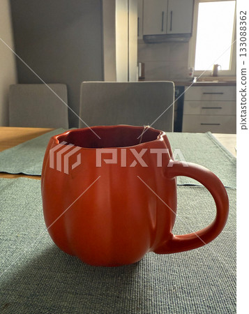 Orange pumpkin shaped mug on dining table in kitchen. Autumn decoration, morning tea, and cozy home lifestyle aesthetic. 133088362