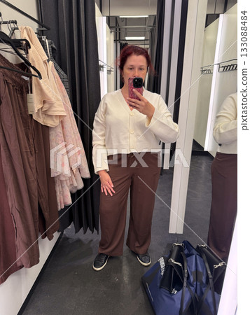 Woman wearing cropped white cardigan with wide brown trousers in fitting room. Relaxed outfit combining softness, simplicity, and balanced neutral tones. 133088484