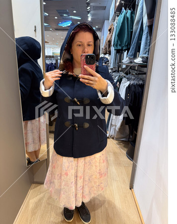 Woman trying on hooded navy duffle coat with toggle closure in fitting room. Warm casual style symbolizing coziness, simplicity, and seasonal charm. Woman trying on hooded navy duffle coat with toggle closure in fitting room. Warm casual style symbolizing coziness, simplicity, and seasonal charm. 133088485